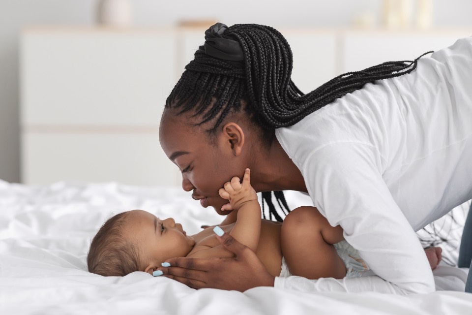 A woman leaning over her baby on a bed, the baby reaching up to touch her face.
